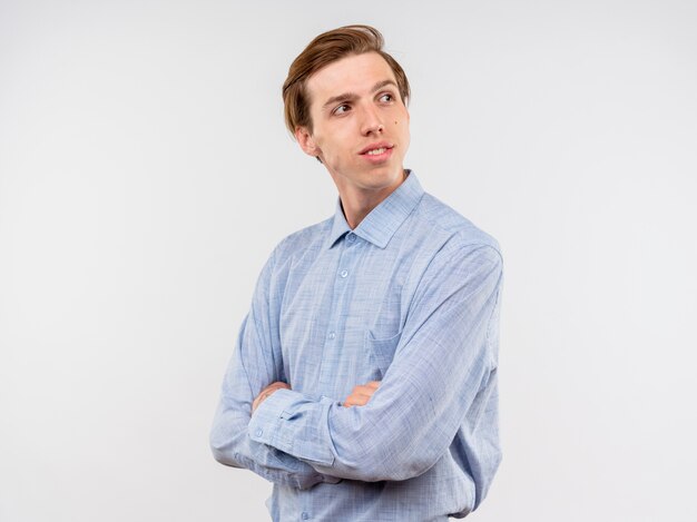 Young man in blue shirt looking aside with serious confident expression standing over white wall