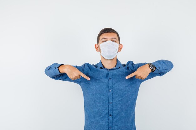 Young man in blue shirt, mask pointing at himself and looking proud , front view.