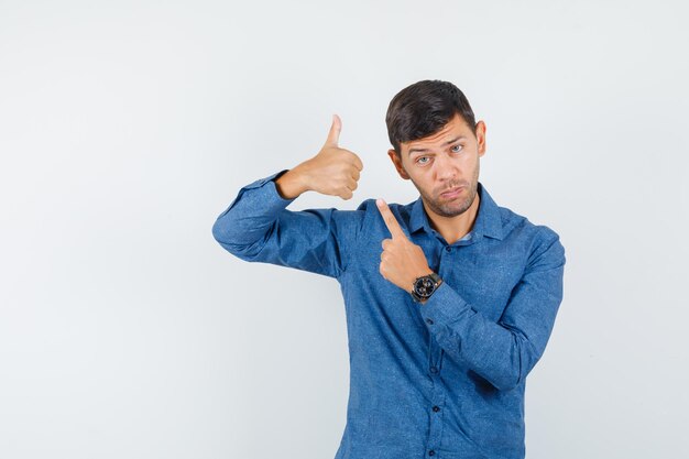 Young man in blue shirt pointing at his thumb up , front view.