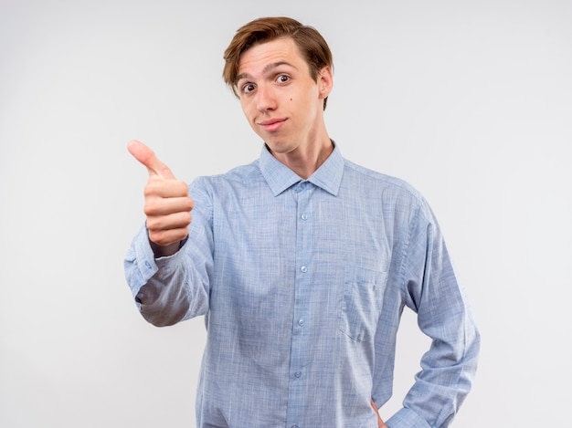 Foto gratuita giovane uomo in camicia blu sorridente che mostra i pollici in su in piedi sul muro bianco