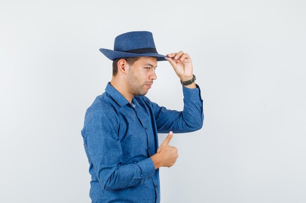 Young man in blue shirt touching his hat and showing thumb up and looking handsome .