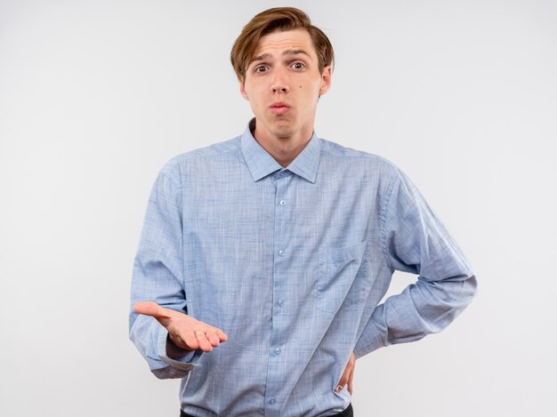 Young man in blue shirt  with arm out being confused and displeased standing over white wall