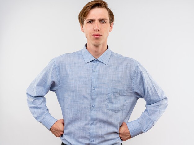 Молодой человек в голубой рубашке с серьезным уверенным выражением лица с руками на бедрах стоит над белой стеной