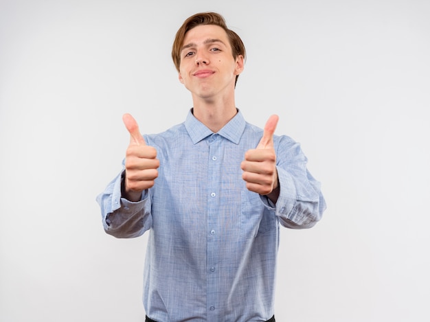 Young man in blue shirt  with smile on face showing thumbs up standing over white wall