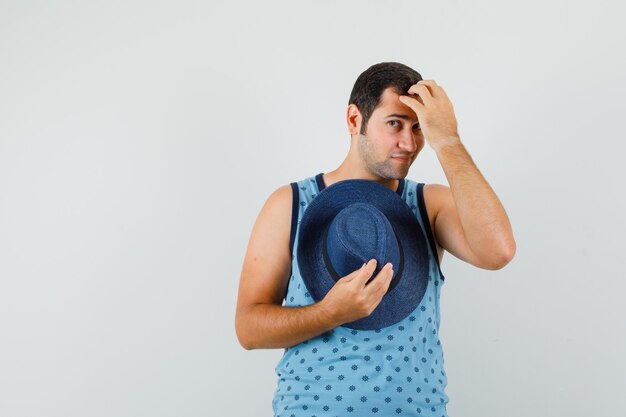 Young man in blue singlet holding hat, combing hair with fingers and looking elegant