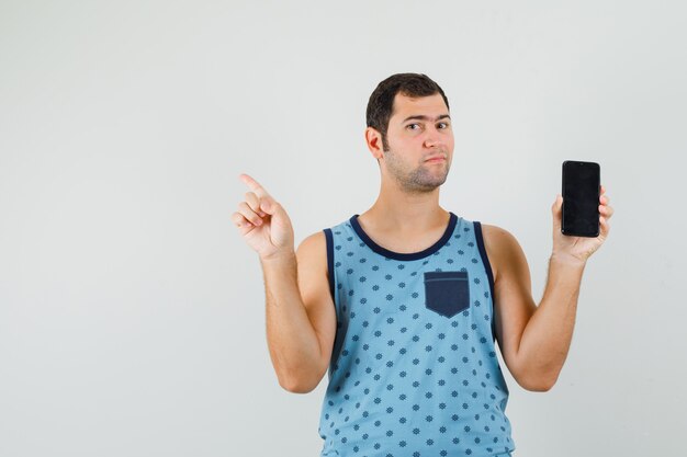 Young man in blue singlet holding mobile phone, pointing away and looking confident