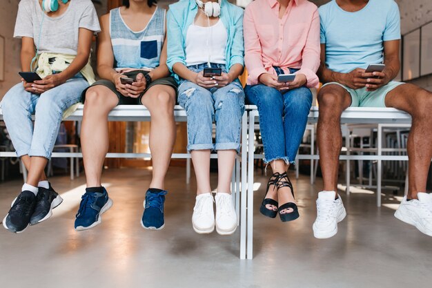 Young man in blue sneakers spending time with friends and typing new message on his cell. Indoor portrait of students sitting together on desk and using smartphones..