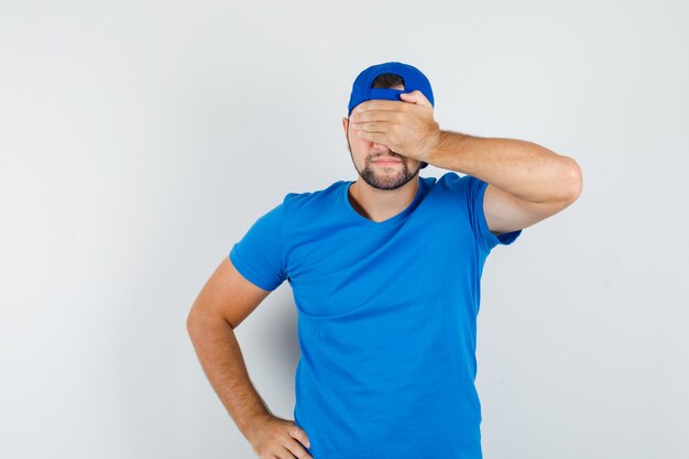 Young man in blue t-shirt and cap covering eyes with hand and looking excited