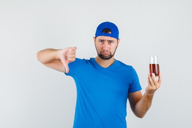 Young man in blue t-shirt and cap holding cold drink with thumb down and looking disappointed