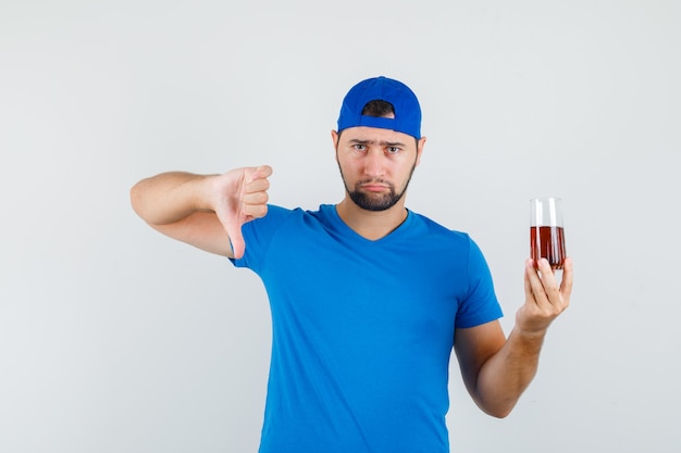 青いTシャツとキャップの若い男は親指を下に向けて冷たい飲み物を保持し、失望しているように見える