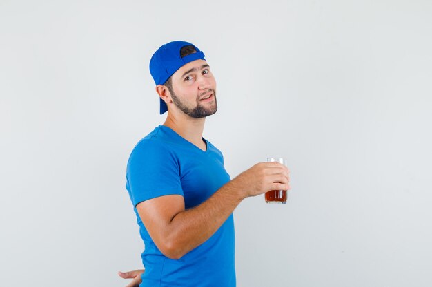 청량 음료 잔을 들고 긍정적 인 찾고 파란색 티셔츠와 모자에 젊은 남자