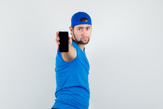 Young man in blue t-shirt and cap holding mobile phone and looking serious