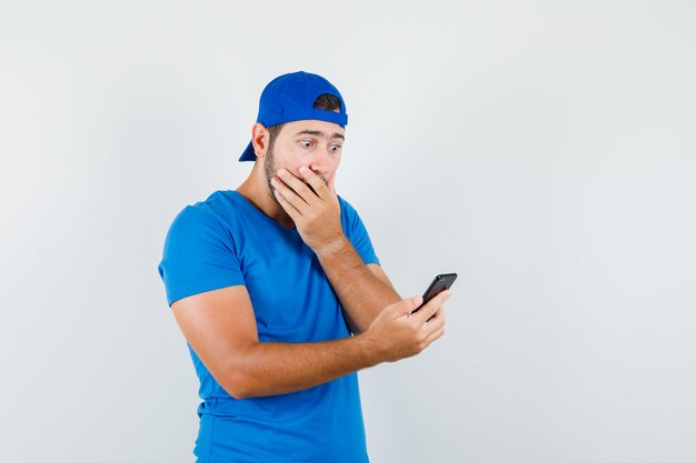 Young man in blue t-shirt and cap looking at mobile phone and looking surprised