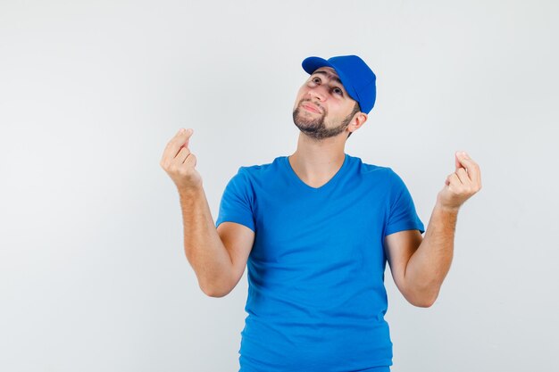 Молодой человек в синей футболке и кепке смотрит вверх, жестикулируя пальцами и выглядит обнадеживающим