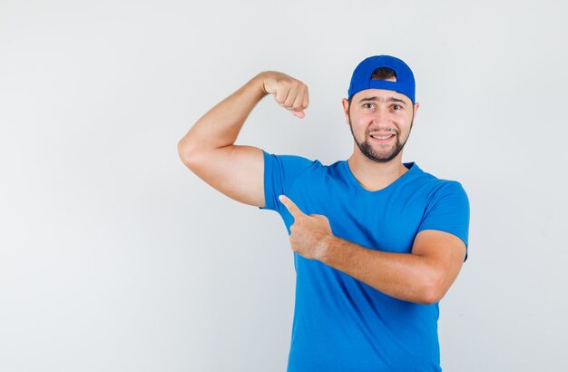 Young man in blue t-shirt and cap pointing at his muscles and looking confident