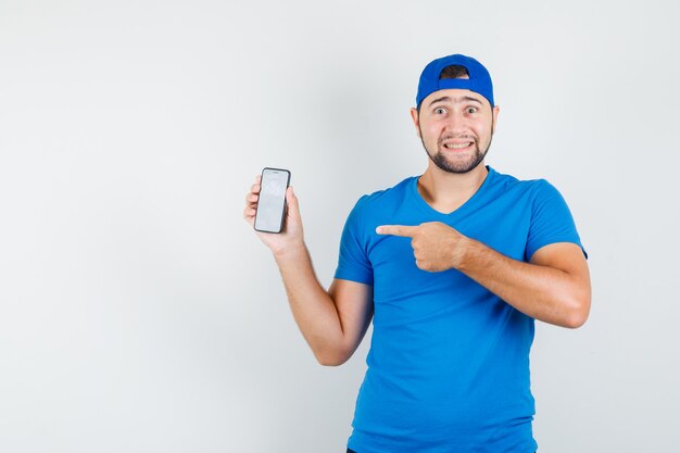 Молодой человек в синей футболке и кепке указывает на мобильный телефон и выглядит веселым