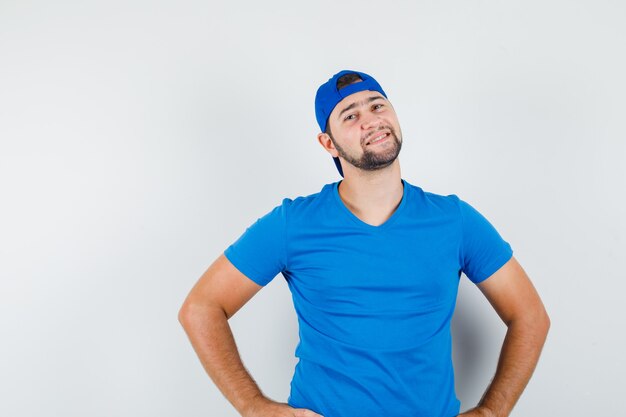 Young man in blue t-shirt and cap standing with hands on waist and looking positive