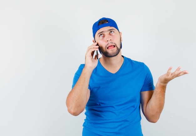 Молодой человек в синей футболке и кепке разговаривает по мобильному телефону и выглядит нерешительно