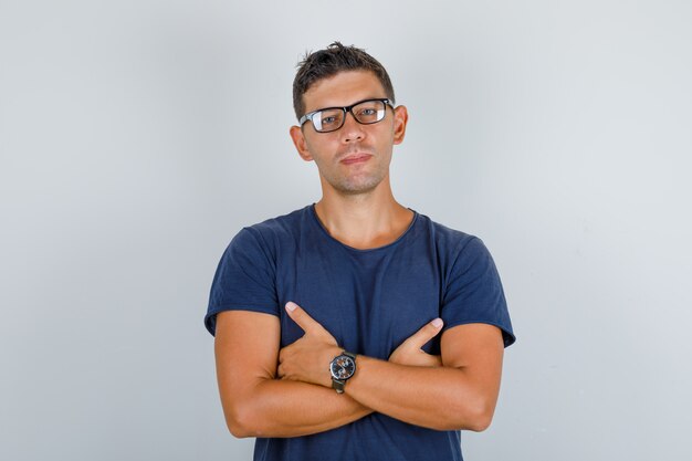 Young man in blue t-shirt, glasses folding hands on chest and looking serious, front view.