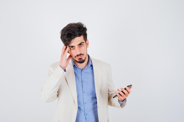 Young man in blue t-shirt and white suit jacket holding one hand to face and holding smartphone and looking serious