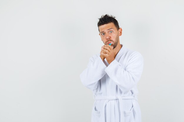 Young man brushing teeth in white bathrobe and looking funny , front view.