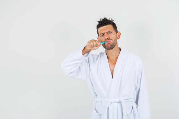 Young man brushing teeth with frowned eyebrows in white bathrobe front view.