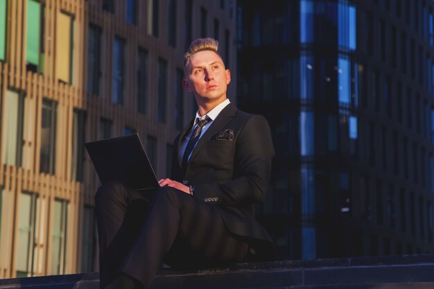 Молодой человек бизнесмен в стильной одежде в костюме сидит на ступеньках с ноутбуком вечером на закате после работы