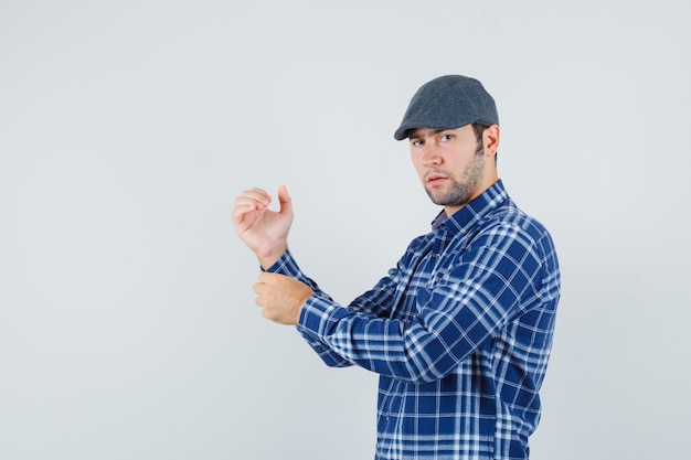 Young man buttoning sleeve of his shirt in shirt, cap and looking pensive. front view.