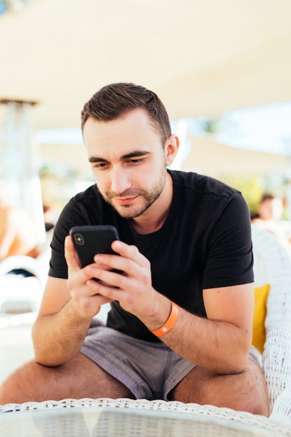 ビーチの夏のリゾートの屋外カフェで携帯電話で呼び出す若い男