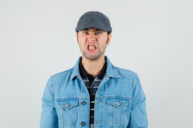 Young man in cap, t-shirt, jacket clenching teeth and looking angry