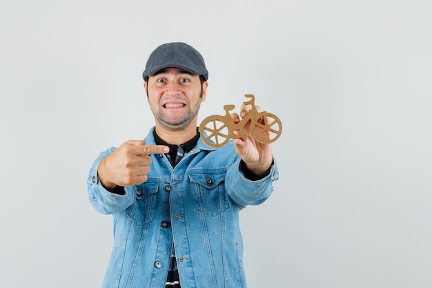 Молодой человек в кепке, футболке, куртке указывает на деревянный игрушечный велосипед и выглядит веселым