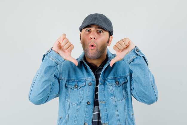 Young man in cap, t-shirt, jacket showing thumbs down and looking surprised