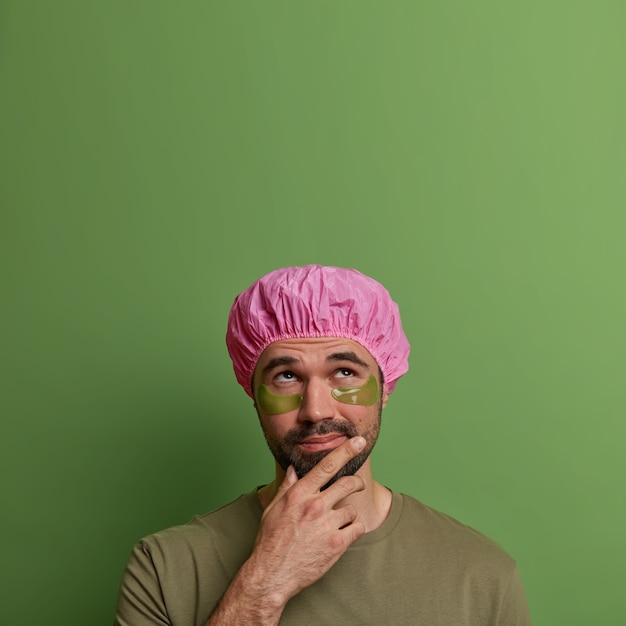 Young man cares about his appearance, reduces wrinkles, applies patches under eyes after taking shower, holds chin and looks thoughtfully above, isolated on green wall, empty space for text