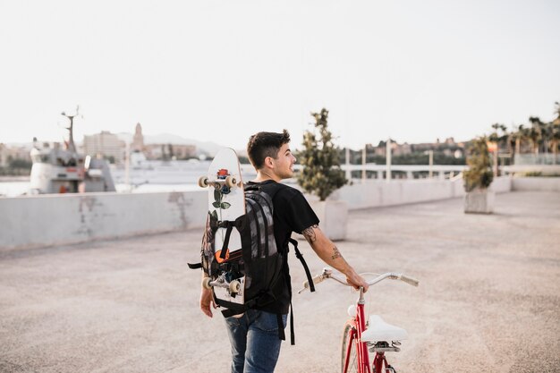 Young man carrying skateboard walking with bicycle