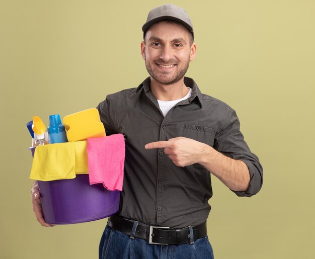 Young man in casual clothes and cap holding bucket with cleaning tools pointing with index finger at it smiling confident standing over green wall