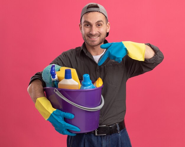 Young man in casual clothes and cap in rubber gloves holding bucket with cleaning tools pointing with index finger at tools happy and positive smiling standing over pink wall