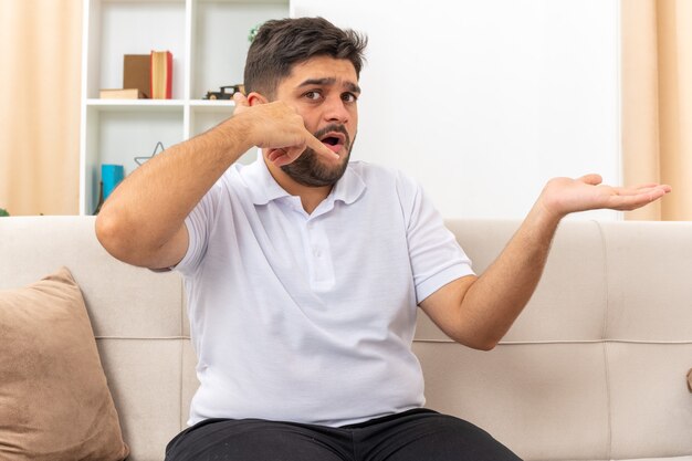 Young man in casual clothes  confused making call me gesture presenting something with arm of hand sitting on a couch in light living room