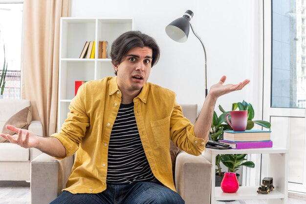 Young man in casual clothes  confused spreading arms to the sides sitting on the chair in light living room