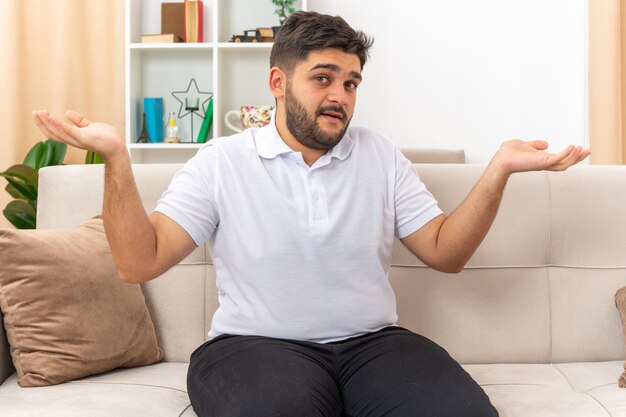 Young man in casual clothes  confused spreading arms to the sides sitting on a couch in light living room