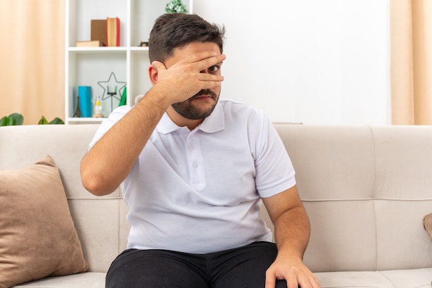 Giovane uomo in abiti casual che copre gli occhi con il palmo guardando attraverso le dita confuso e preoccupato che trascorre il fine settimana a casa seduto su un divano in un soggiorno luminoso