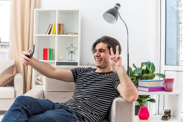 Молодой человек в повседневной одежде делает селфи с помощью смартфона, показывая v-знак счастливым и веселым, сидя на стуле в светлой гостиной