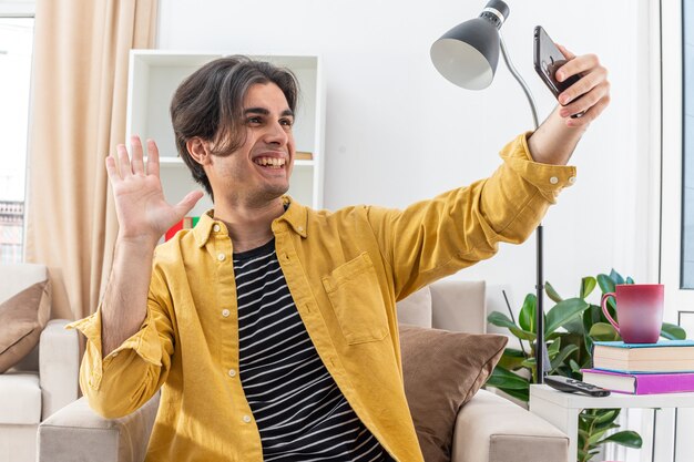 Young man in casual clothes doing selfie using smartphone waving with hand happy and cheerful smiling broadly sitting on the chair in light living room