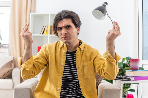 Young man in casual clothes gesturing with hands in displeasure and indignation sitting on the chair in light living room