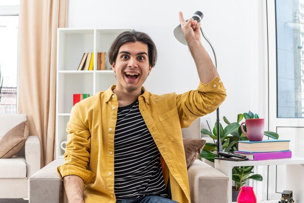 Young man in casual clothes  happy and surprised showing index finger having new great idea sitting on the chair in light living room