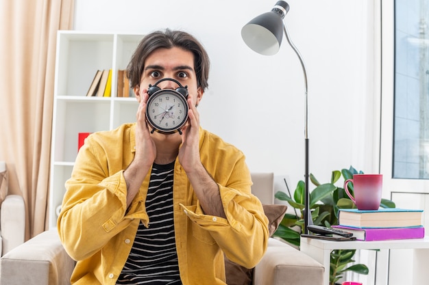 Free photo young man in casual clothes holding alarm clock in front of his face looking worried sitting on the chair in light living room