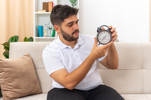 Young man in casual clothes holding alarm clock looking at it with serious face sitting on a couch in light living room