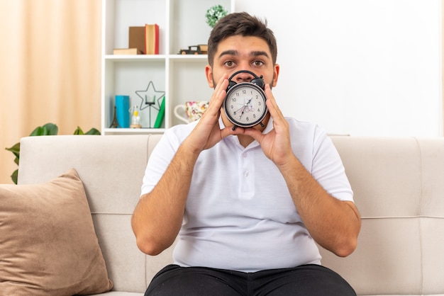 Young man in casual clothes holding alarm clock looking  worried sitting on a couch in light living room