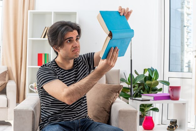 Young man in casual clothes holding book looking at it with sad expression sitting on the chair in light living room