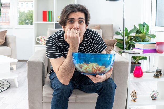 Young man in casual clothes holding bowl of chips looking tv stressed and nervous biting nails sitting on the chair in light living room