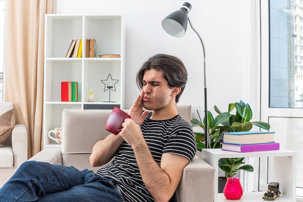 Young man in casual clothes holding cup of tea looking unwell touching his cheek feeling toothache sitting on the chair in light living room